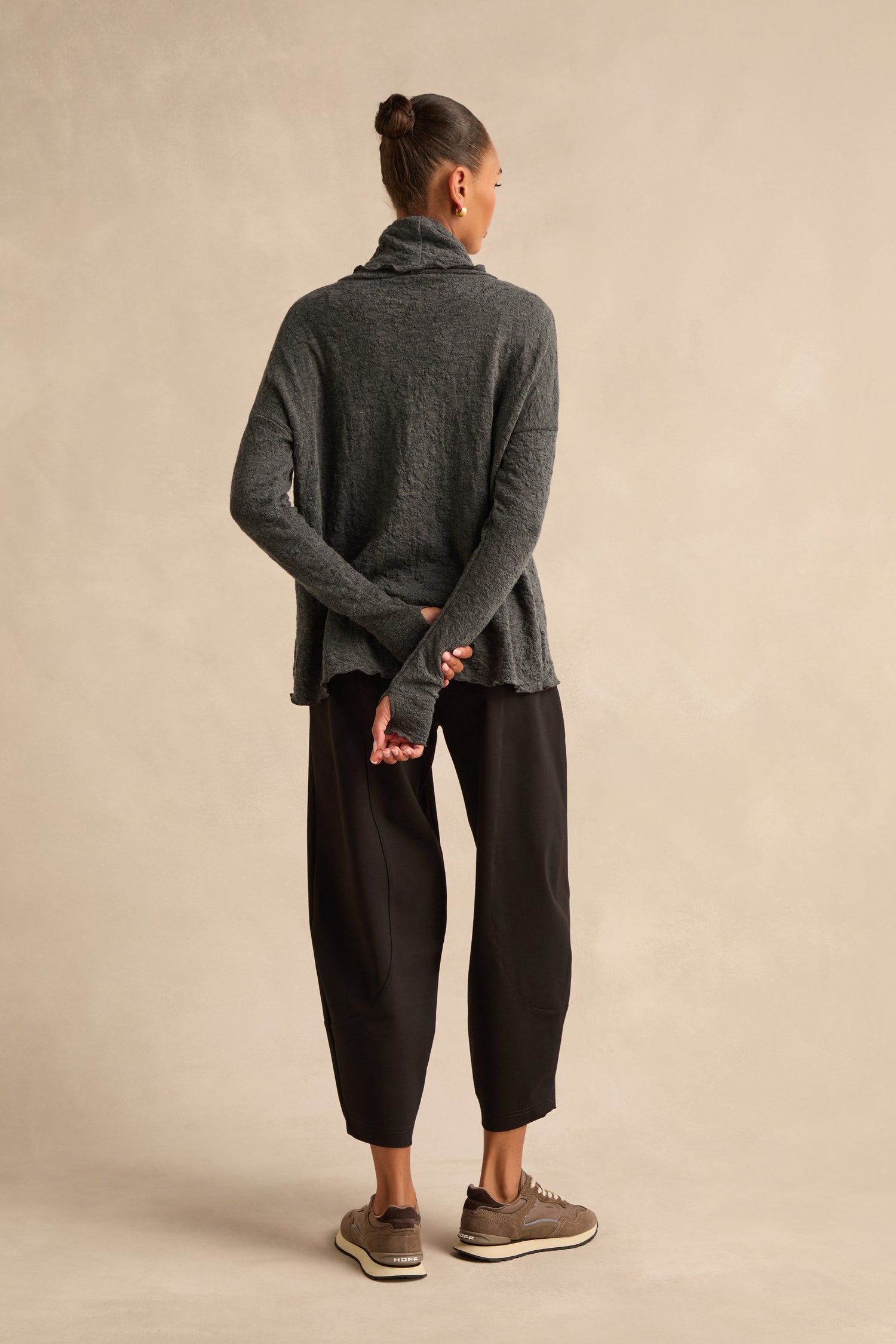 A person stands facing away, hands clasped behind their back, wearing the Anthracite Textured Merino Roll Neck Top, black loose pants, and brown sneakers against a beige background.