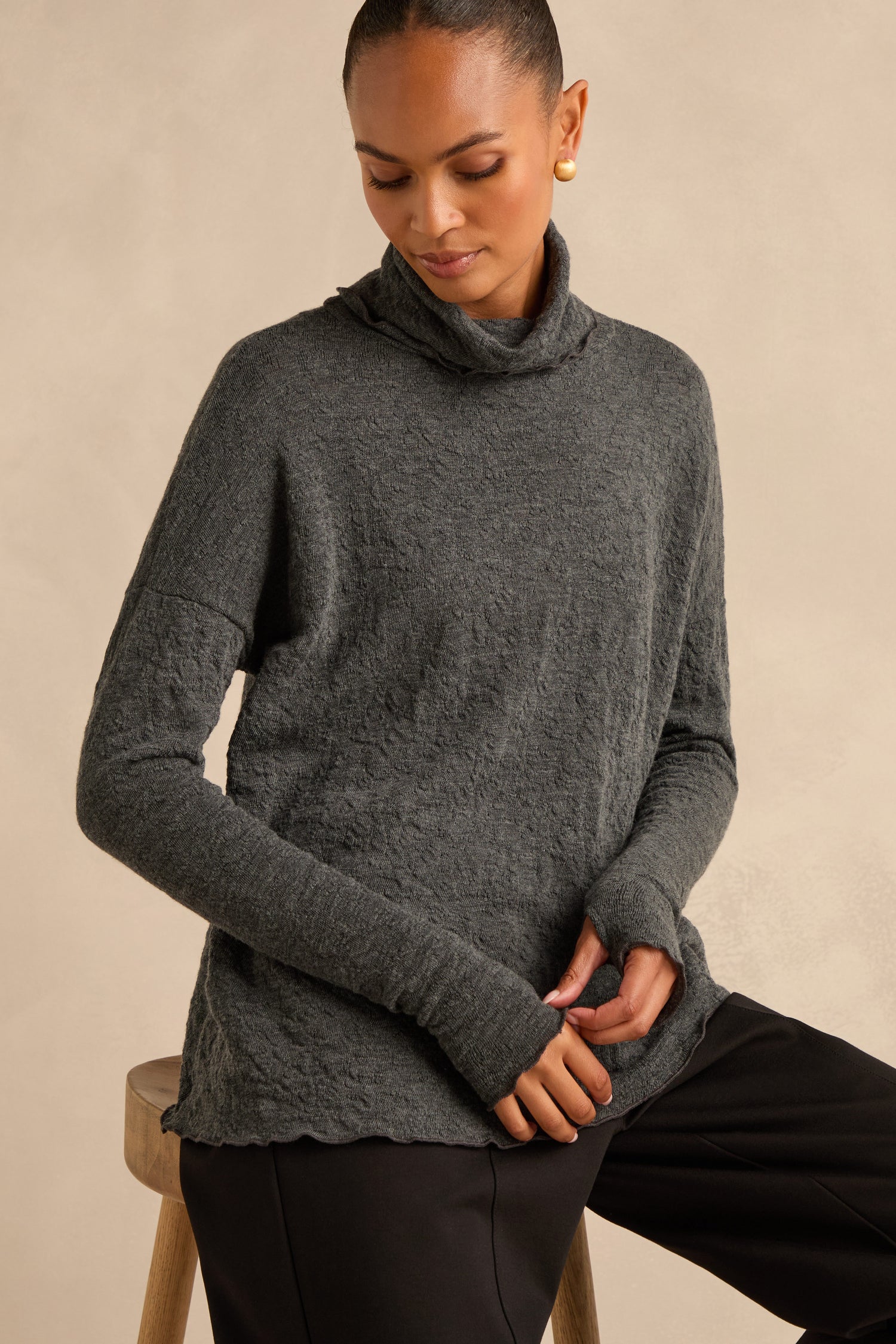 A person sits on a stool wearing the Anthracite Textured Merino Roll Neck Top—crafted in Denmark—paired with black pants and gold earrings, their hair pulled back and hands resting in their lap.