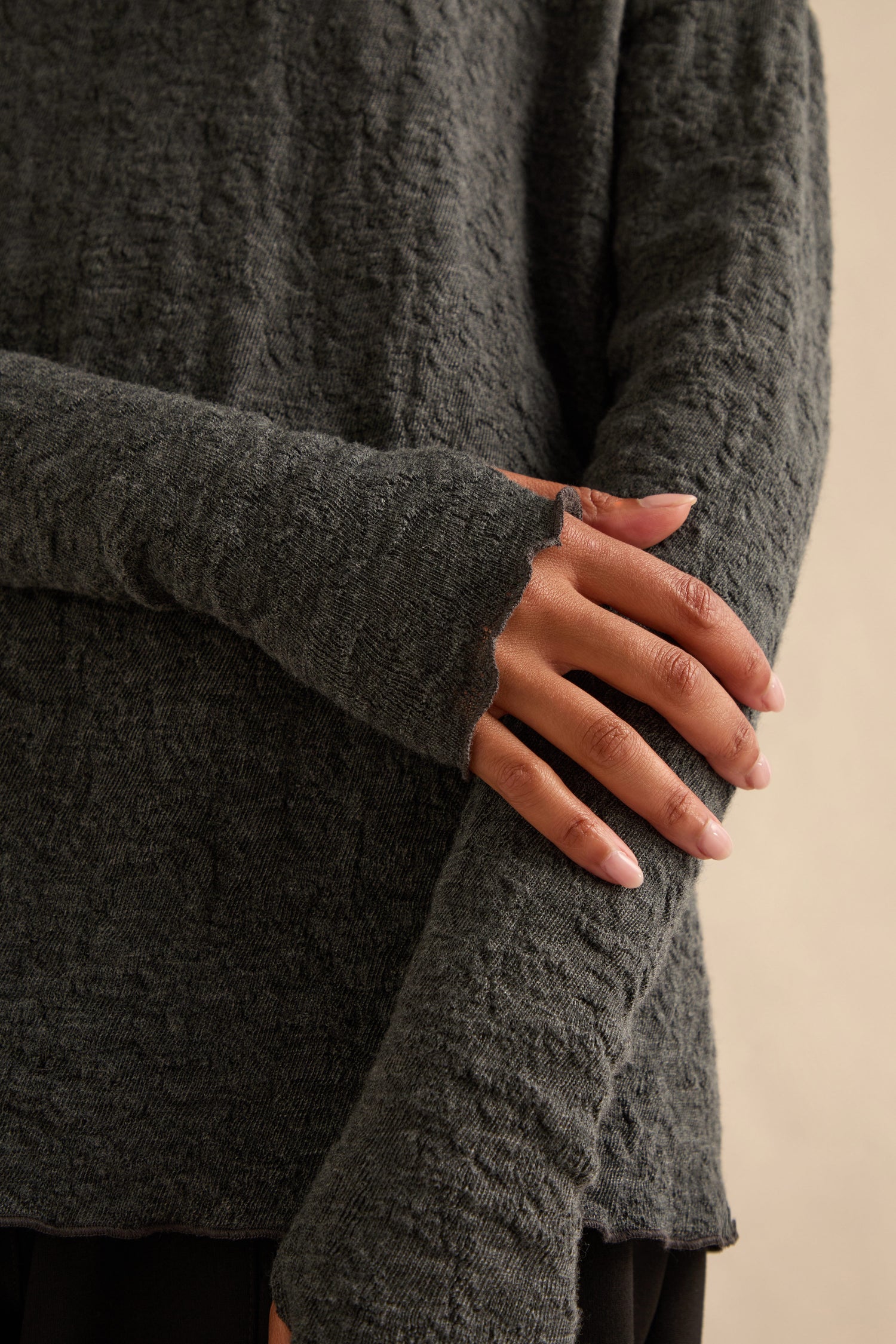 Wearing the Anthracite Textured Merino Roll Neck Top—a long-sleeve, dark grey knit—the person poses with one arm crossed over their body, displaying natural nails and a relaxed posture.