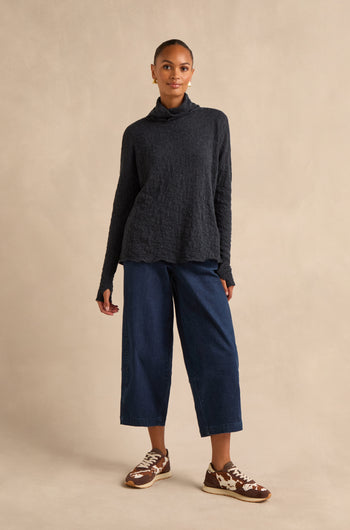 A person stands against a beige background wearing a Navy Textured Merino Roll Neck Top, wide-leg blue jeans, and brown sneakers.