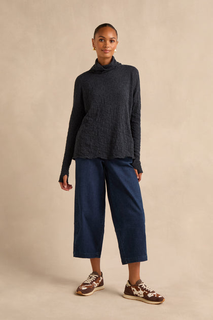 A person stands against a beige background wearing a Navy Textured Merino Roll Neck Top, wide-leg blue jeans, and brown sneakers.
