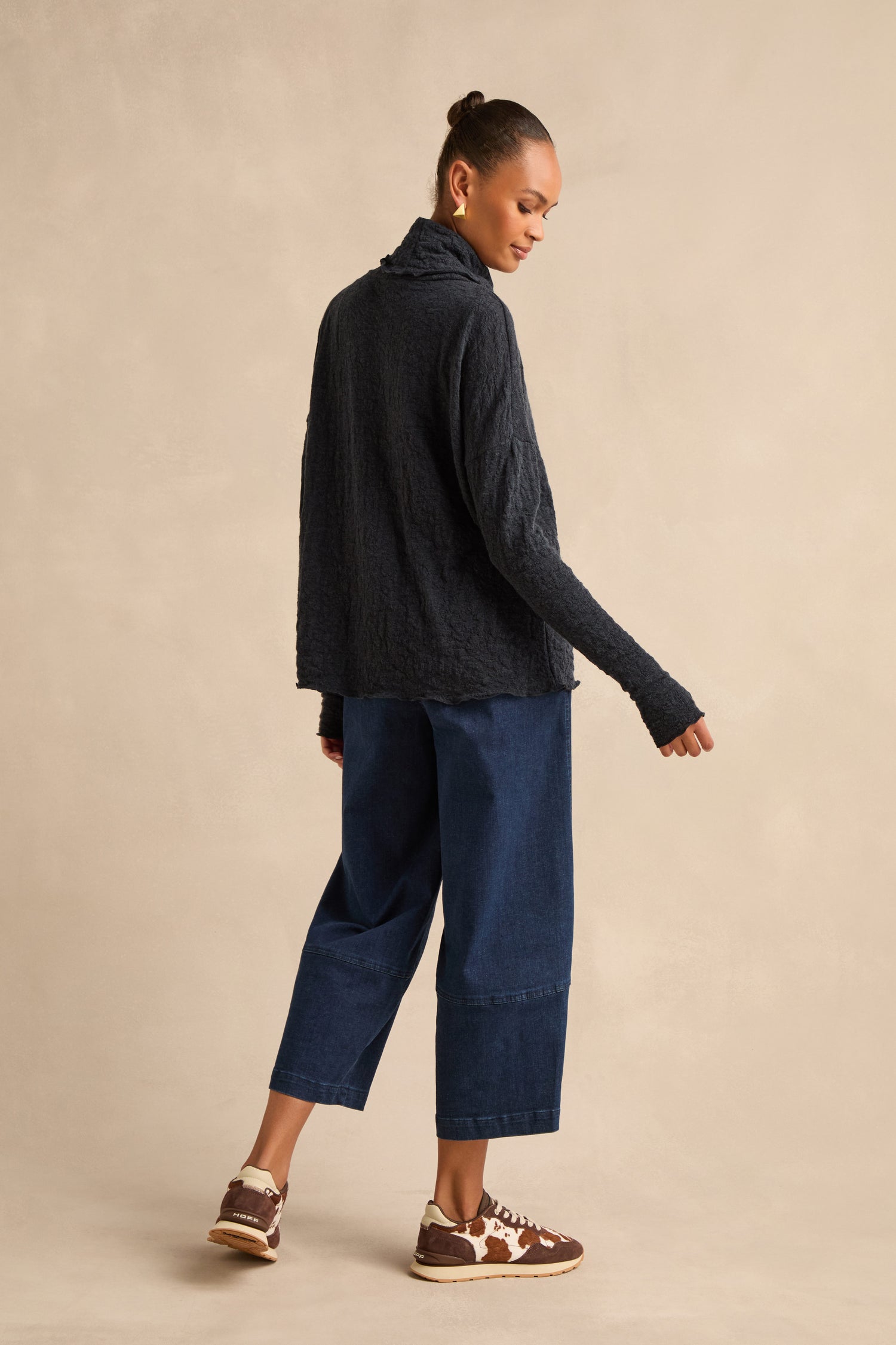 A woman stands facing slightly away, wearing the Navy Textured Merino Roll Neck Top with wide-leg blue jeans and brown sneakers, set against a plain beige background.