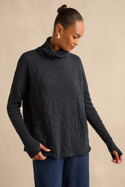 A woman wears the Navy Textured Merino Roll Neck Top with blue jeans, standing against a neutral background and looking down.