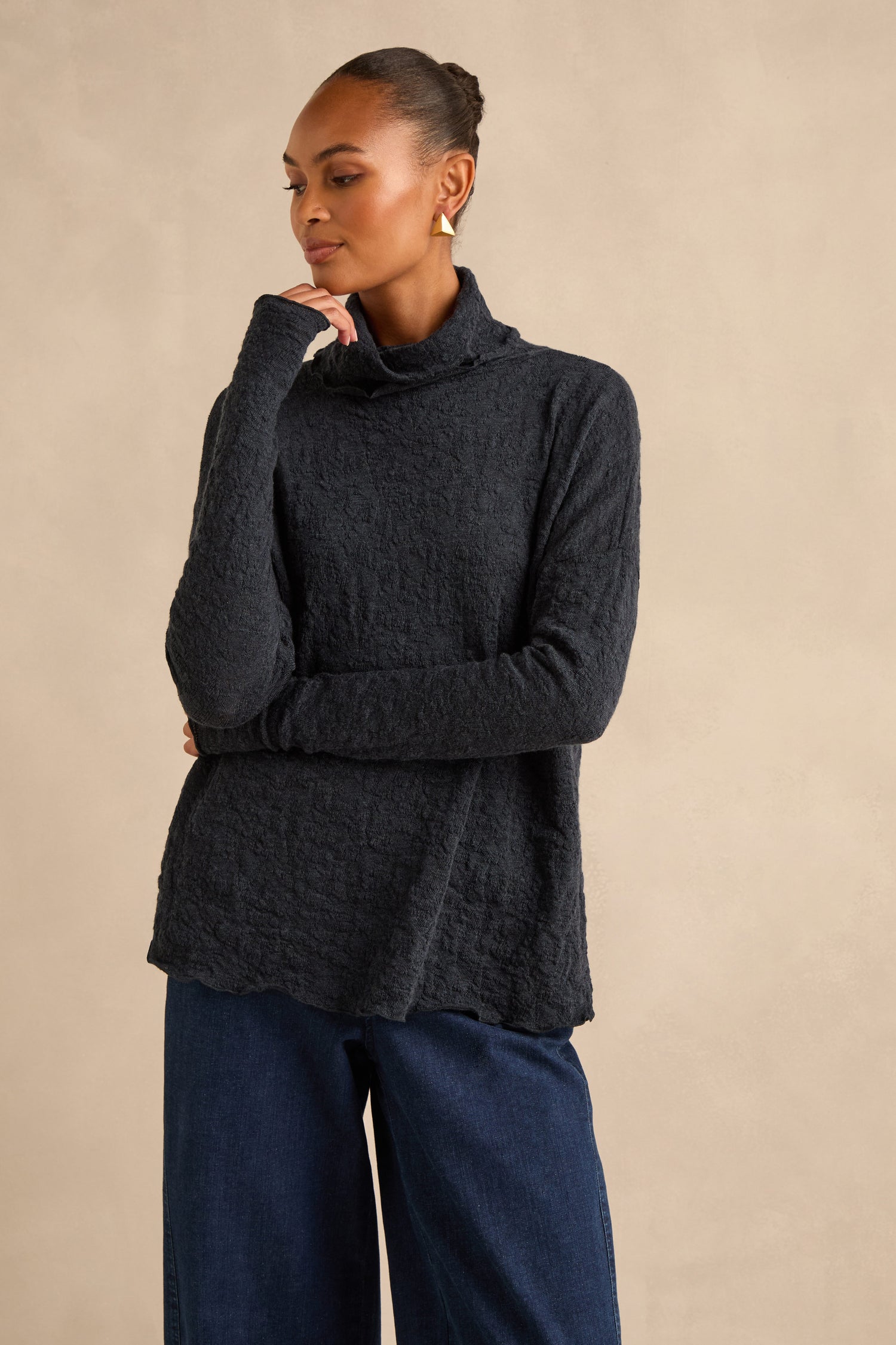 A woman wearing a Navy Textured Merino Roll Neck Top and wide-leg dark blue jeans stands against a plain beige background.