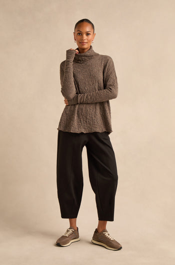A woman stands against a plain background, wearing the made-to-order Taupe Textured Merino Roll Neck Top, paired with loose black pants and brown sneakers, her left hand touching her chin.