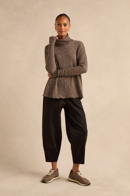 A woman stands against a plain background, wearing the made-to-order Taupe Textured Merino Roll Neck Top, paired with loose black pants and brown sneakers, her left hand touching her chin.