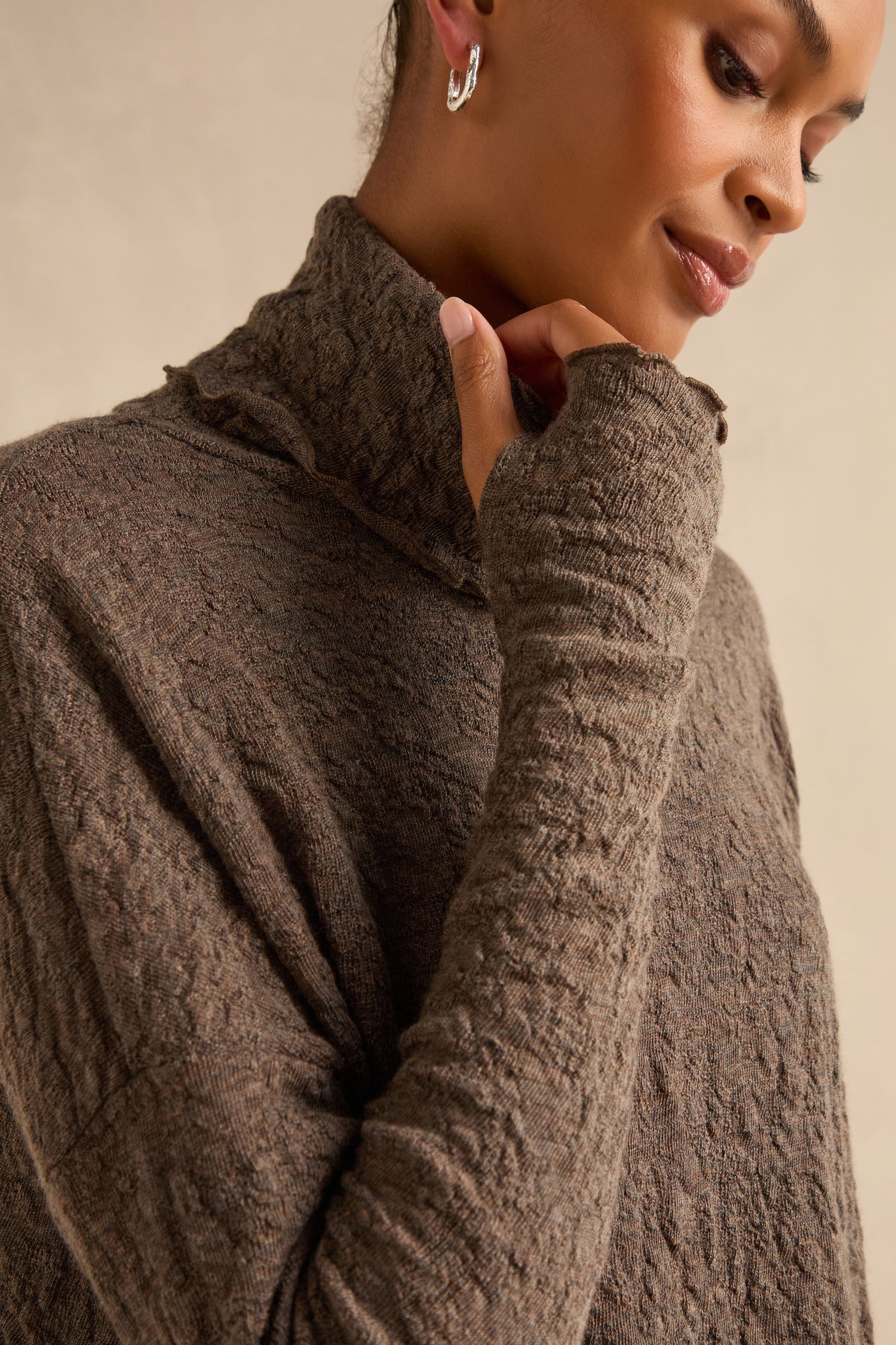 Wearing the Taupe Textured Merino Roll Neck Top with thumbhole sleeves and silver hoop earrings, the person poses against a plain background. This made-to-order piece features a unique textured knit in merino wool for added warmth and style.