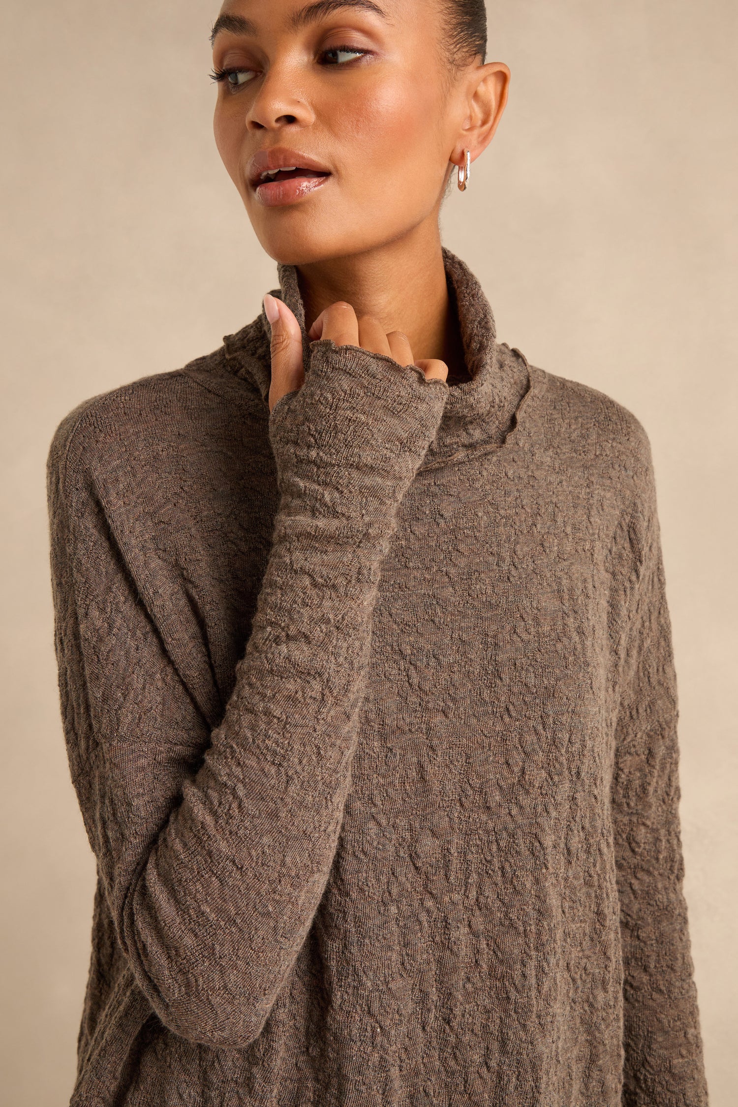 A person wearing the Taupe Textured Merino Roll Neck Top stands against a plain beige background, looking slightly to the side. This merino wool piece exudes understated elegance and cozy comfort.