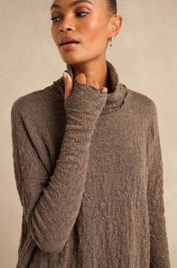 A person wearing the Taupe Textured Merino Roll Neck Top stands against a plain beige background, looking slightly to the side. This merino wool piece exudes understated elegance and cozy comfort.