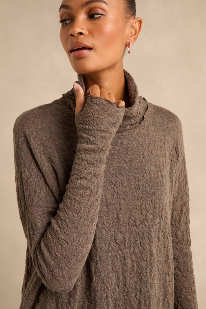 A person wearing the Taupe Textured Merino Roll Neck Top stands against a plain beige background, looking slightly to the side. This merino wool piece exudes understated elegance and cozy comfort.