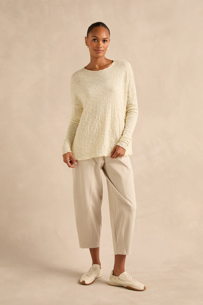 Woman standing against a beige background wearing the Ivory Textured Merino Scoop Neck Top, light beige cropped pants, and white sneakers.