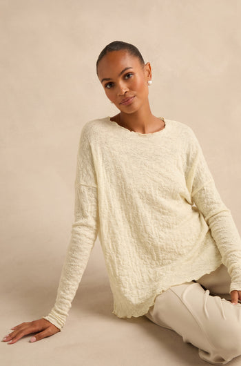 An individual with short hair sits on the floor against a beige background, wearing the Ivory Textured Merino Scoop Neck Top paired with light pants.