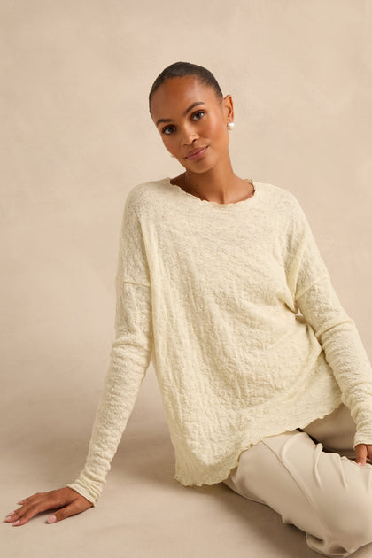 An individual with short hair sits on the floor against a beige background, wearing the Ivory Textured Merino Scoop Neck Top paired with light pants.