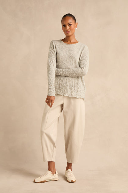 A woman stands against a beige backdrop wearing the Silver Textured Merino Scoop Neck Top, paired with loose cream-colored pants and beige sneakers, her hair pulled back.