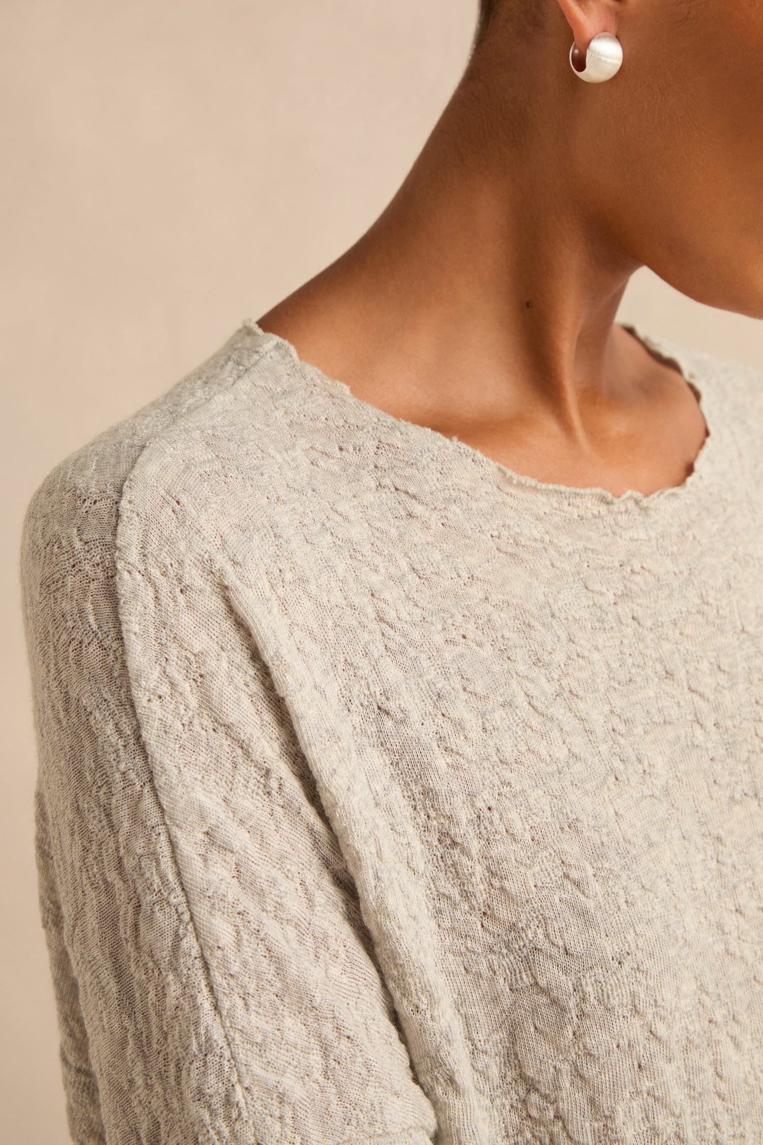 Close-up of a person wearing the Silver Textured Merino Scoop Neck Top—a light gray sweater crafted from merino wool—paired with a pearl earring, highlighting their neck, shoulder, and ear against a neutral background for a refined nod to slow fashion.