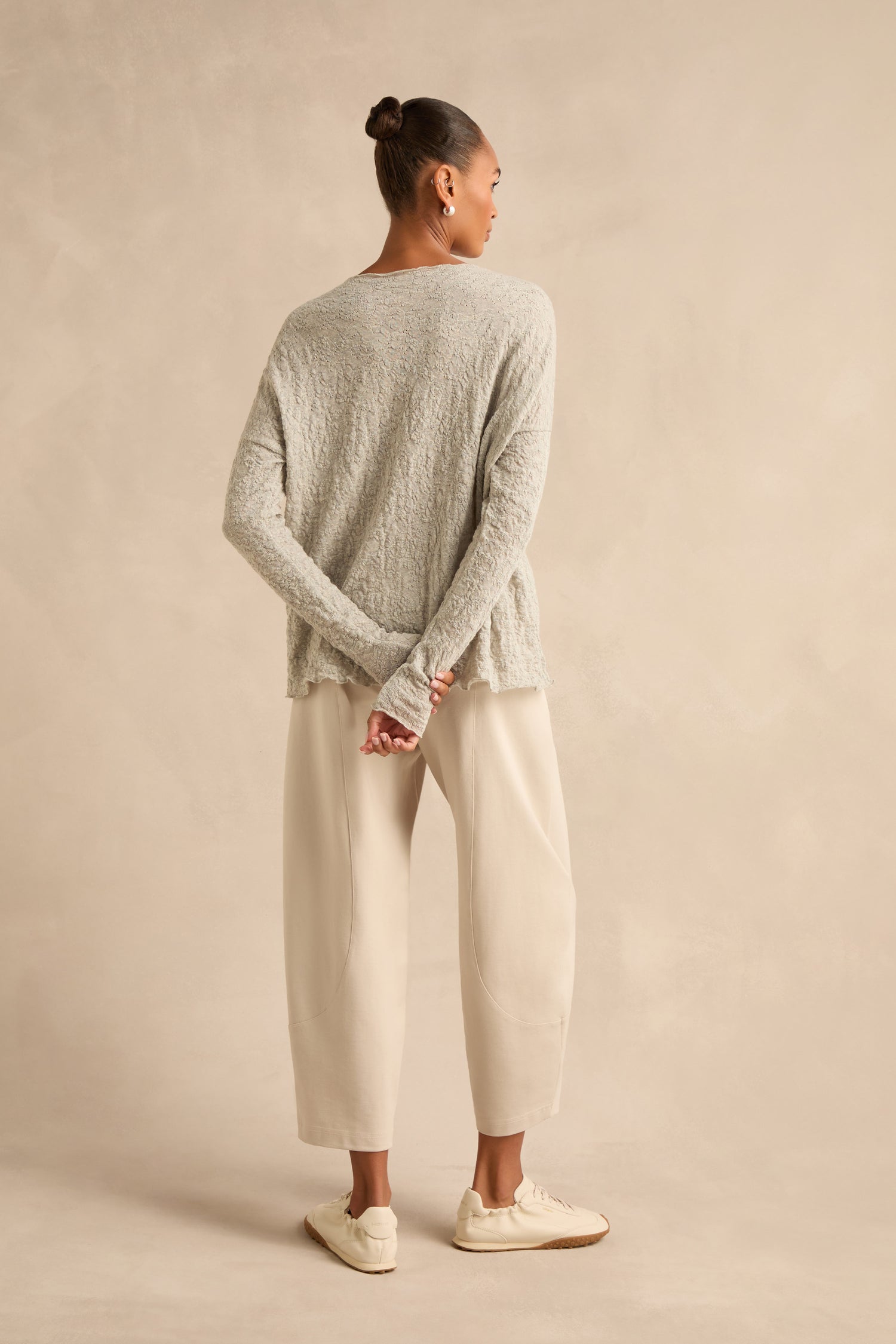 A person stands with hands behind their back, wearing the Silver Textured Merino Scoop Neck Top, cream pants, and white sneakers, facing a beige wall—a nod to timeless slow fashion style.