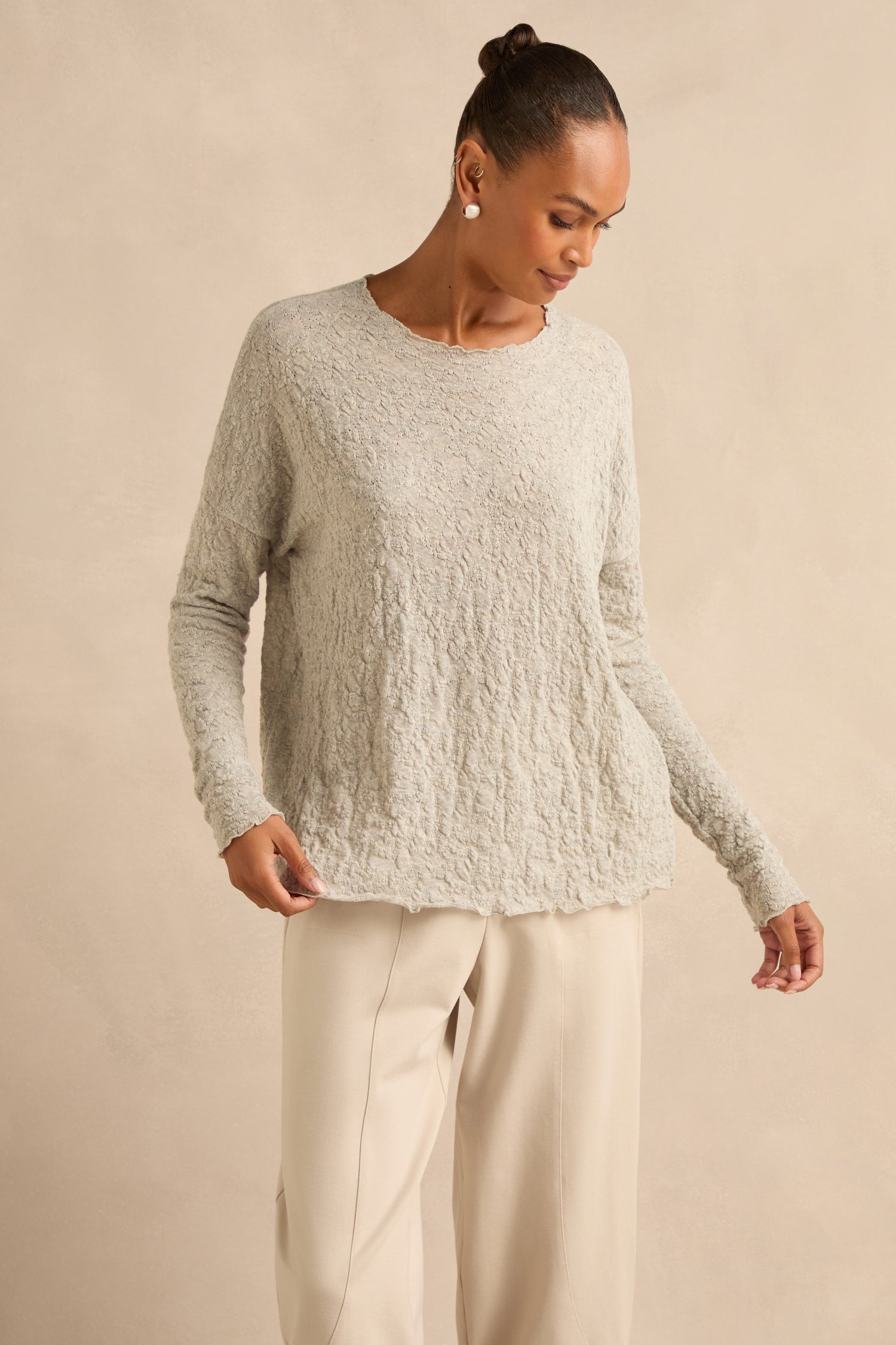 Wearing the Silver Textured Merino Scoop Neck Top and cream pants, a woman stands against a plain beige background, looking down to her left—a timeless look inspired by slow fashion principles.