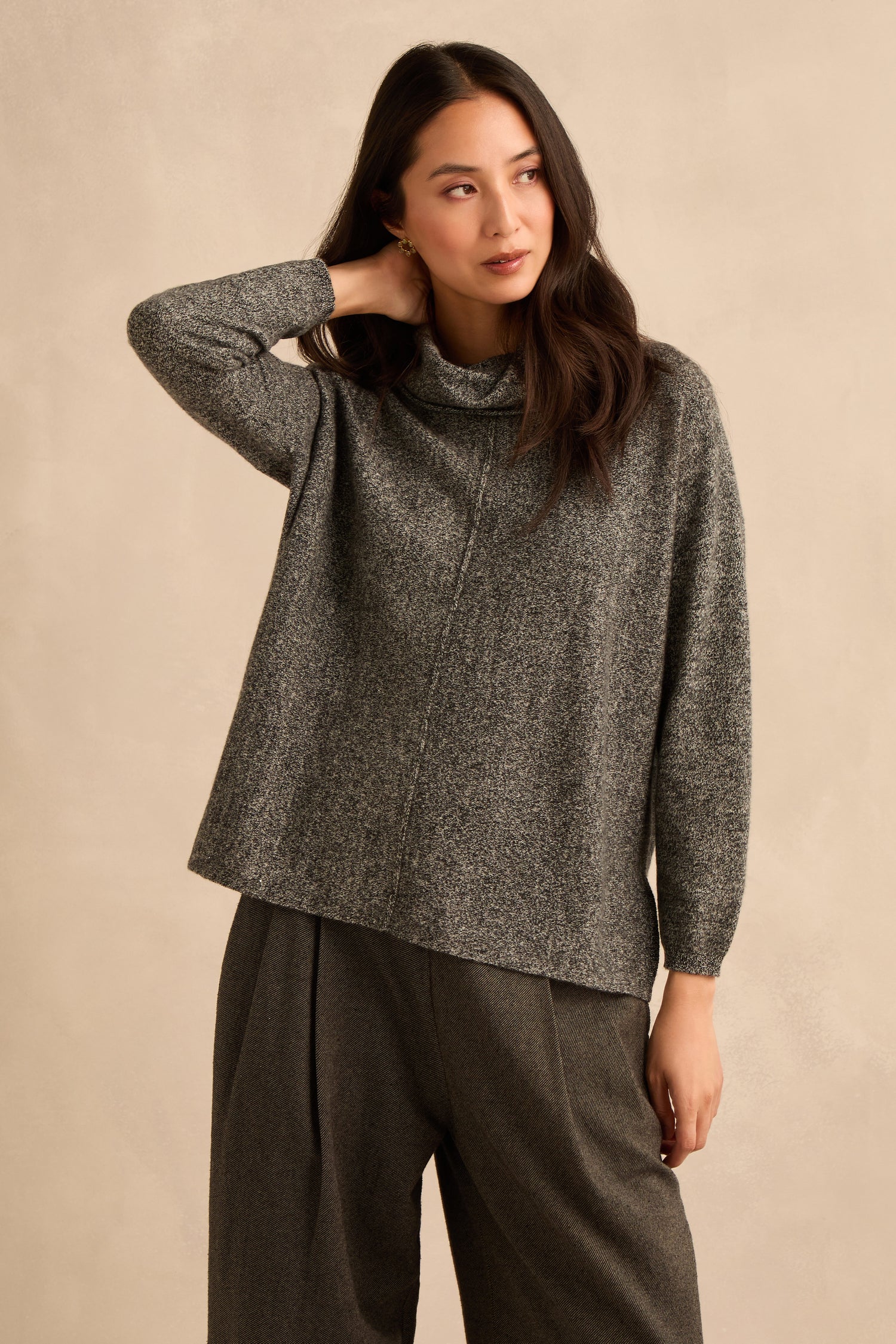 A woman wearing a Black & White Merino Wool Cowl Neck Jumper and wide-leg pants stands against a beige background, touching her neck with one hand and looking to the side.