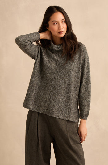 A woman wearing a Black & White Merino Wool Cowl Neck Jumper and wide-leg pants stands against a beige background, touching her neck with one hand and looking to the side.