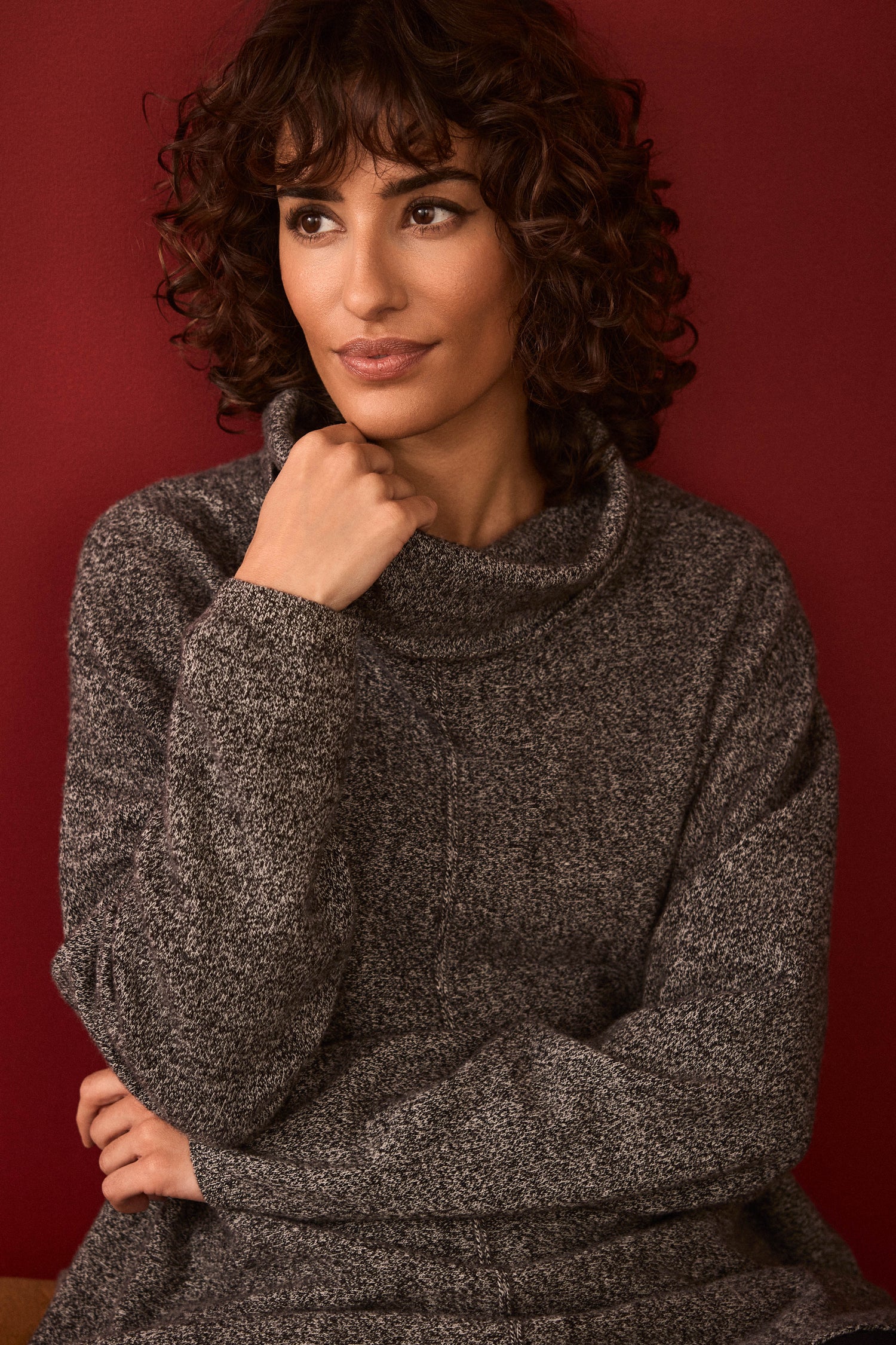 A person with curly hair wears the Black & White Merino Wool Cowl Neck Jumper, sitting with arms crossed in front of a plain red background.