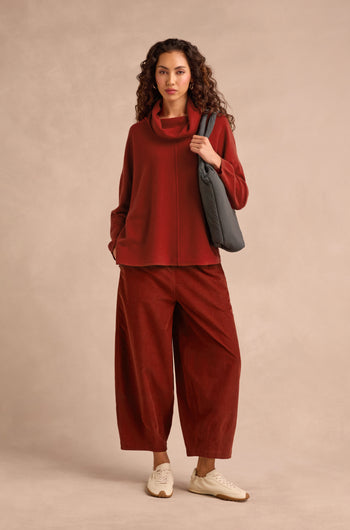 A woman stands against a neutral background wearing the Conker Merino Wool Cowl Neck Jumper in red, paired with matching wide-leg pants, white sneakers, and carrying a black shoulder bag.