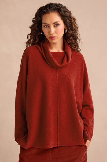 A woman with curly hair wears the Conker Merino Wool Cowl Neck Jumper in a loose-fitting, rust hue, paired with a matching skirt. She stands against a neutral background, showcasing how this cosy knitwear ensemble delivers perfect comfort and effortless style.