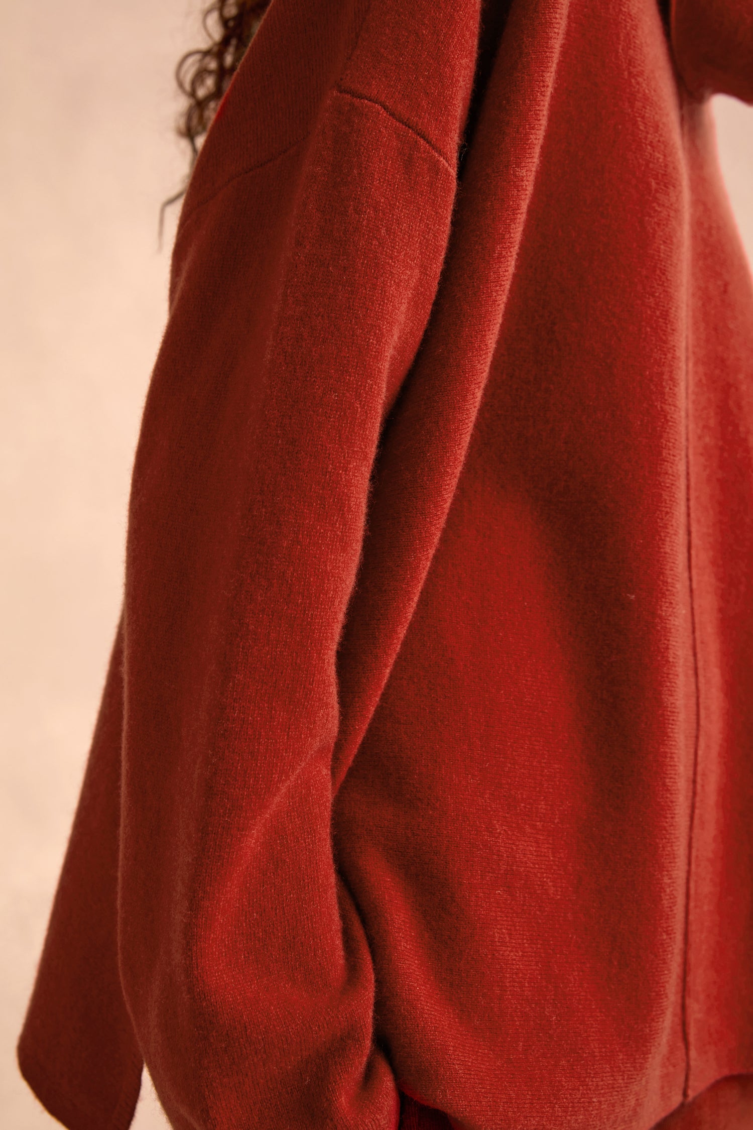 A person models the Conker Merino Wool Cowl Neck Jumper, its relaxed fit and rich red hue highlighted as they are shown from the side, with only part of the upper body and one arm visible. The cosy knitwear stands out against a softly blurred, neutral background.