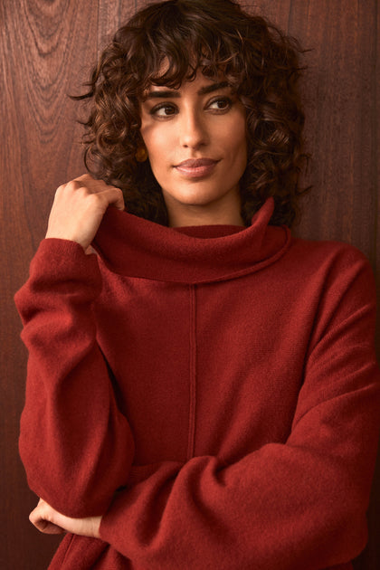 A woman with curly hair stands in front of a wooden wall, wearing the Conker Merino Wool Cowl Neck Jumper. She holds the collar with one hand and gazes slightly to the side.