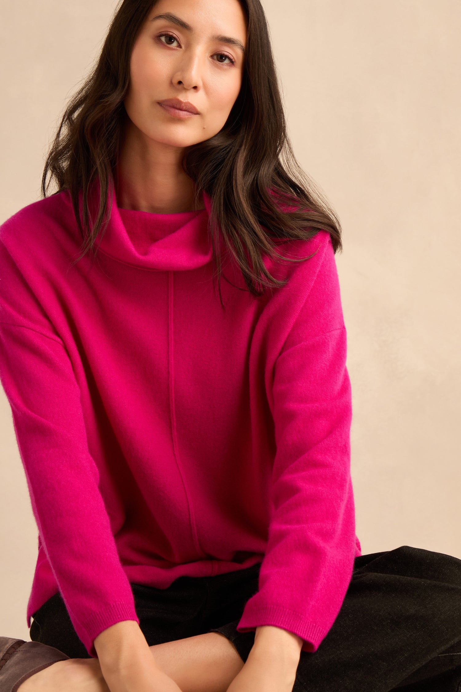 A woman with long dark hair wears the Magenta Merino Wool Cowl Neck Jumper and black pants, seated with arms relaxed against a neutral beige background.