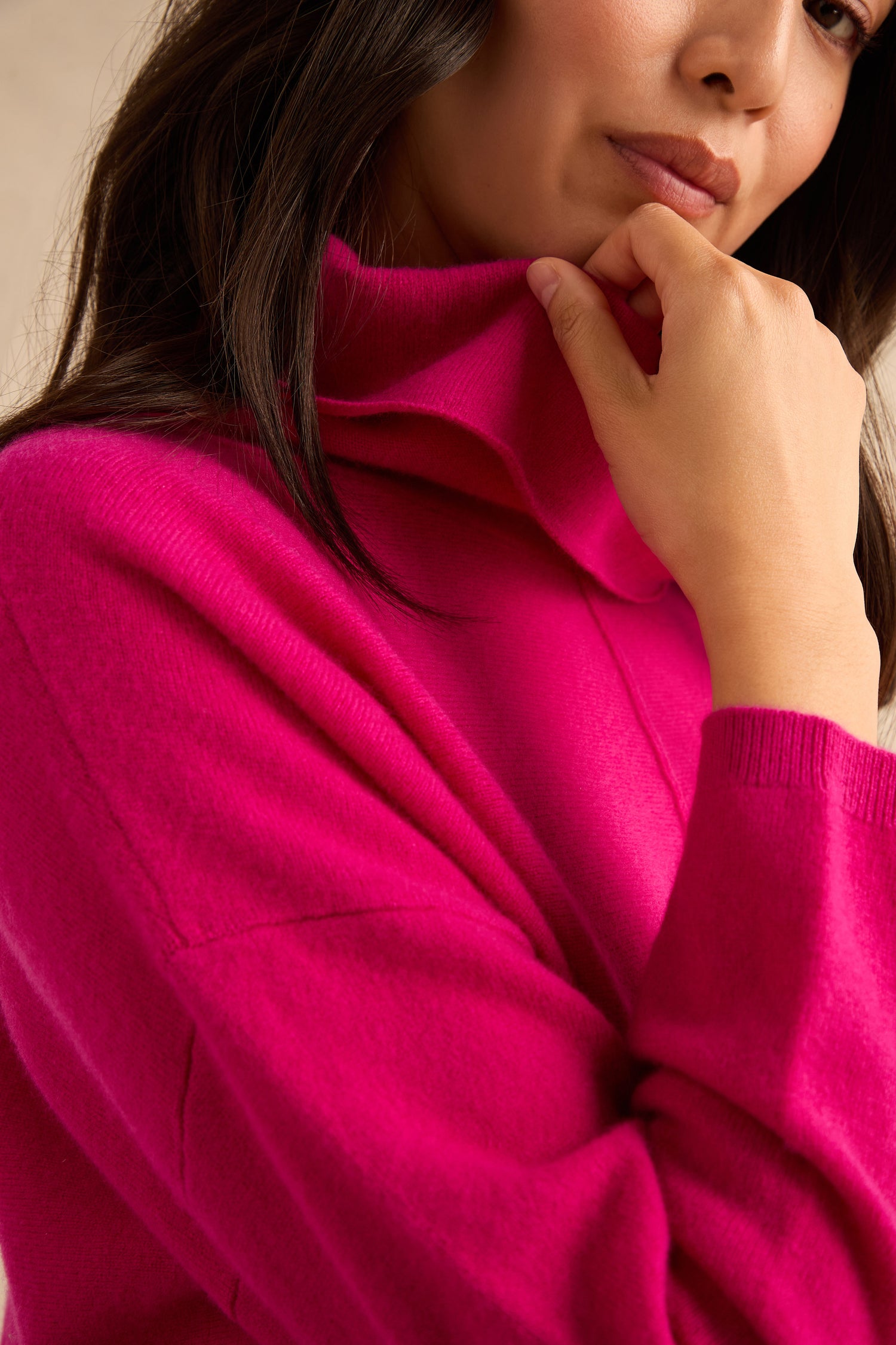 A woman wearing the Magenta Merino Wool Cowl Neck Jumper holds the collar and rests her chin on her hand.