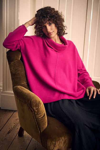 A person wearing a Magenta Merino Wool Cowl Neck Jumper and a black skirt, sitting in a velvet armchair.