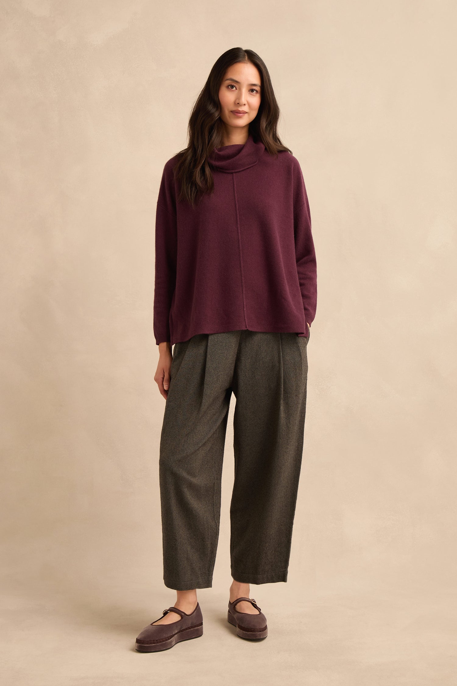 A woman stands facing the camera, wearing the Plum Merino Wool Cowl Neck Jumper, paired with wide gray pants and matching purple shoes, against a beige background.