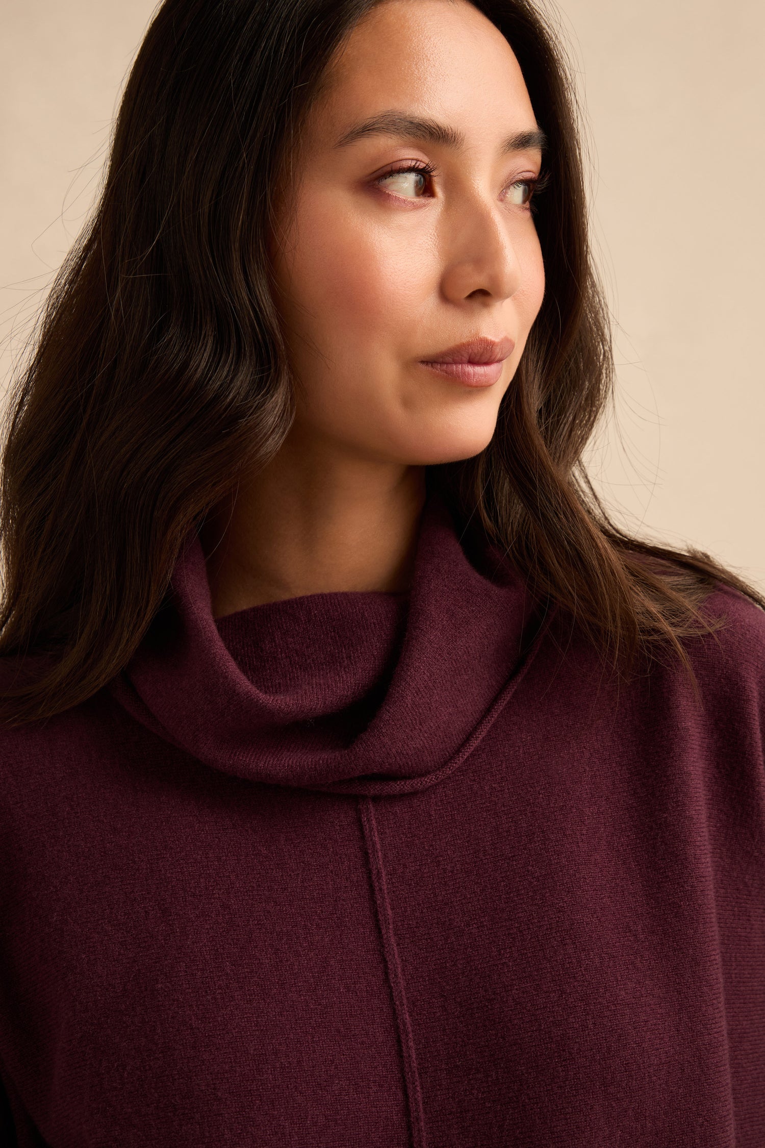 A woman with long dark hair wears the Plum Merino Wool Cowl Neck Jumper, a cosy knit in rich burgundy, as she looks to the side against a neutral background.