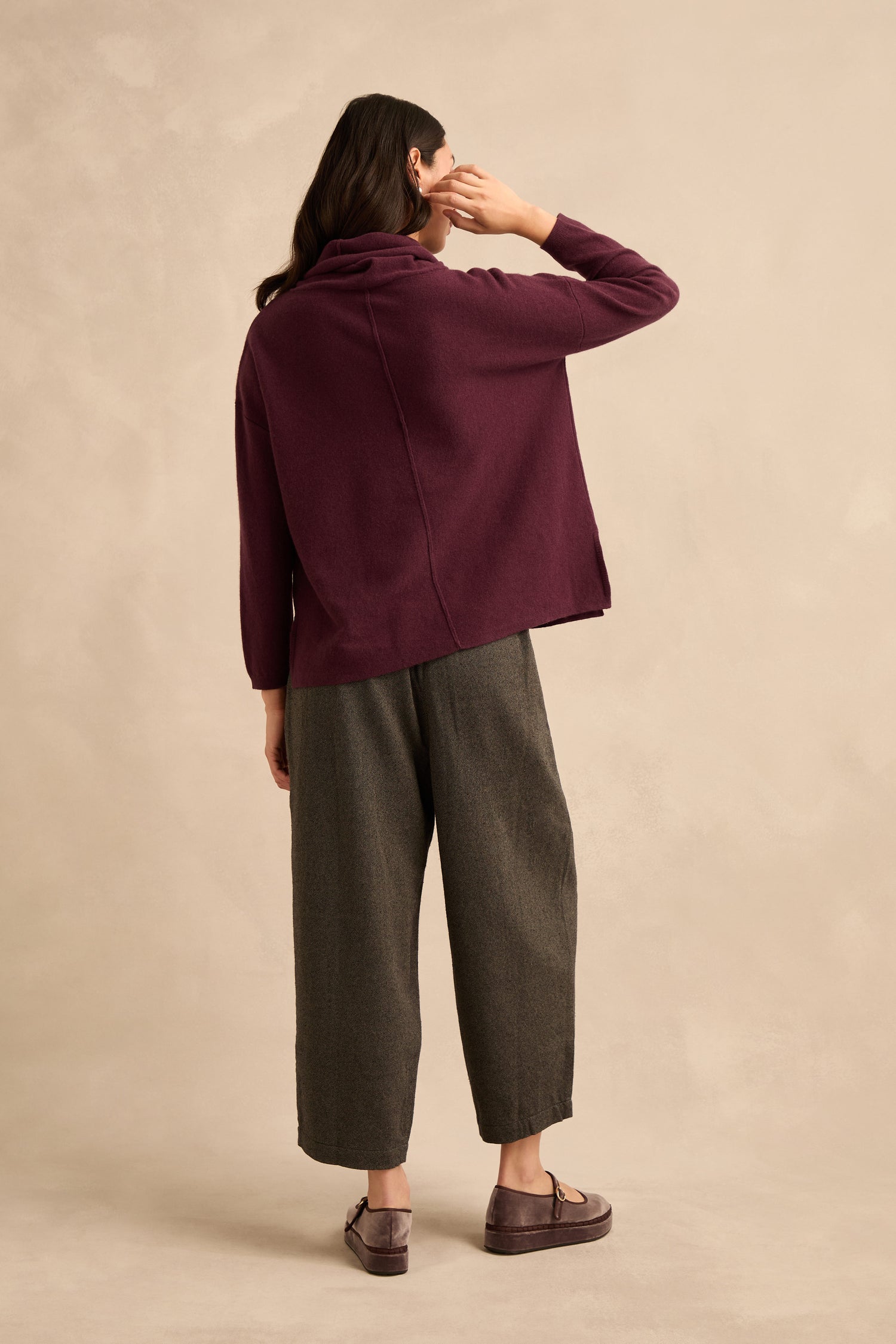 A person stands facing away, wearing the Plum Merino Wool Cowl Neck Jumper, wide gray pants, and mauve loafers, against a beige studio backdrop.
