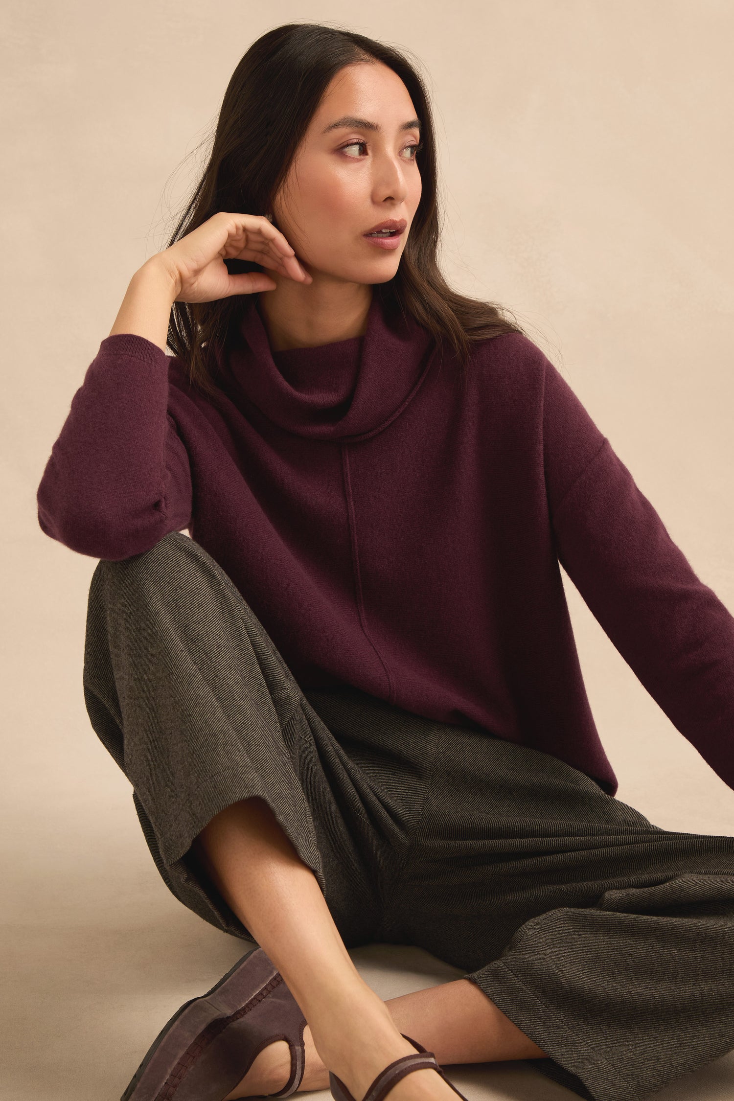 A woman sits on the floor wearing the Plum Merino Wool Cowl Neck Jumper, paired with gray wide-leg pants and brown flats. She looks to her left with one hand resting on her face.