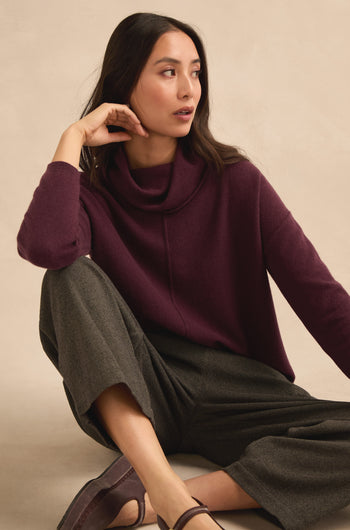 A woman sits on the floor wearing the Plum Merino Wool Cowl Neck Jumper, paired with gray wide-leg pants and brown flats. She looks to her left with one hand resting on her face.
