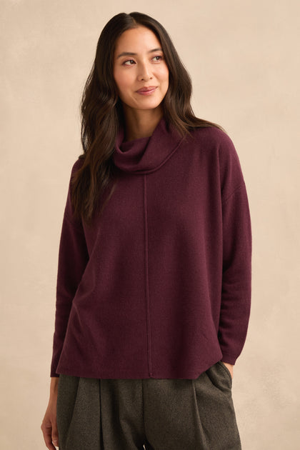 A woman wearing a Plum Merino Wool Cowl Neck Jumper and gray trousers stands against a beige background, gazing slightly to the side.