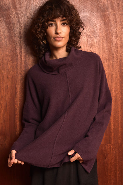A woman with curly hair stands against a wooden wall, holding the hem of her Plum Merino Wool Cowl Neck Jumper and looking at the camera.