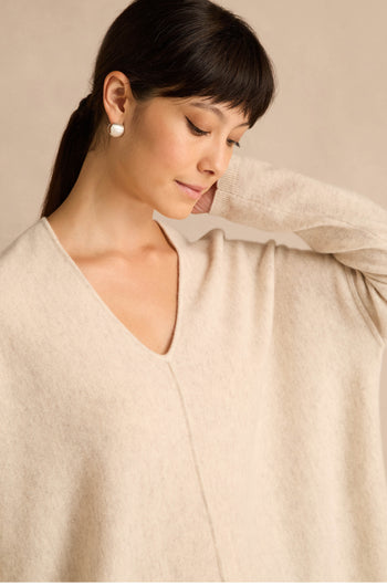 A woman with dark hair and pearl earrings wears an Ecru Merino Wool V Neck Jumper, looking down with one hand gently touching the back of her neck.