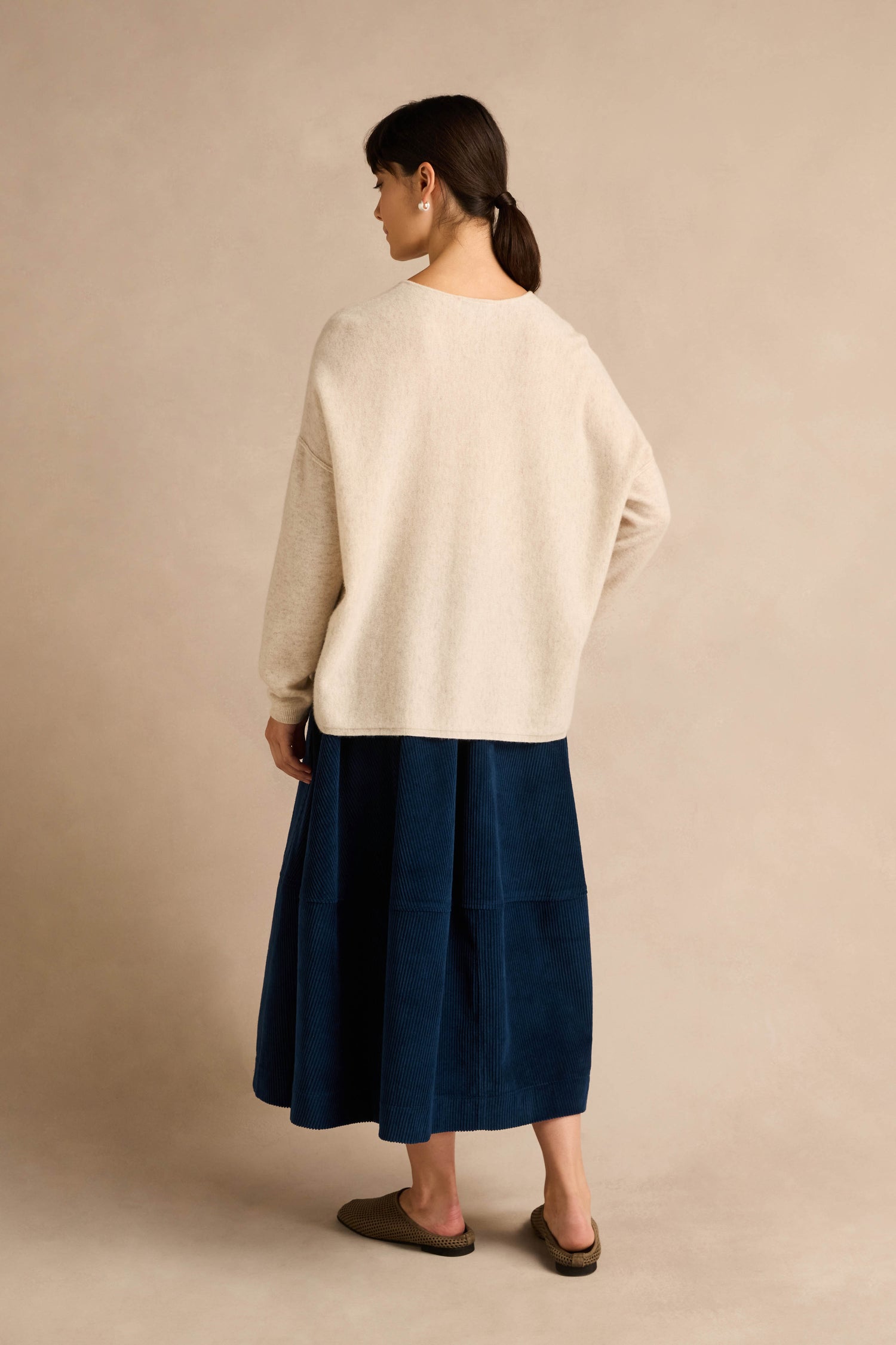 A woman stands facing away, wearing the Ecru Merino Wool V Neck Jumper, a blue midi skirt, and brown slip-on shoes, against a neutral beige background.