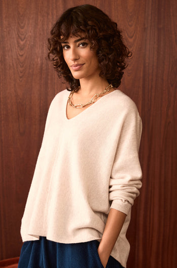 A woman with curly hair wears the Ecru Merino Wool V Neck Jumper, blue pants, and a gold chain necklace as she stands in front of a wooden wall, looking at the camera with a slight smile.