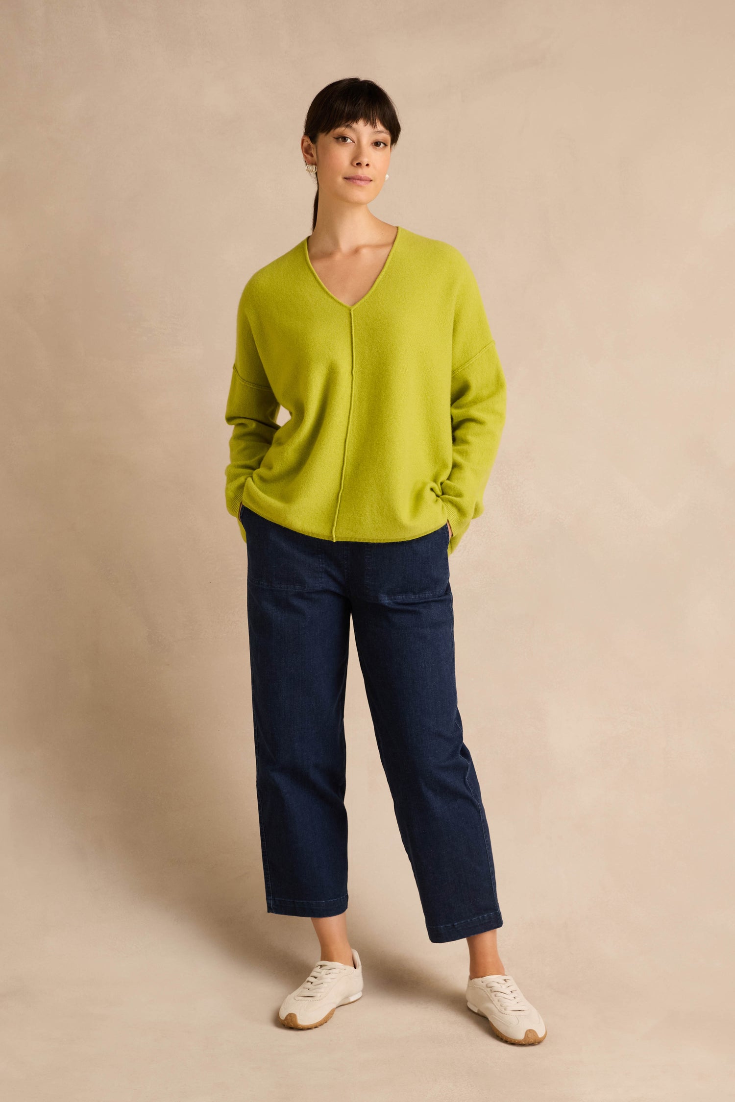 A person stands against a neutral background wearing the Kiwi Merino Wool V Neck Jumper in lime green, paired with dark blue jeans and white sneakers, with their hands in their pockets.