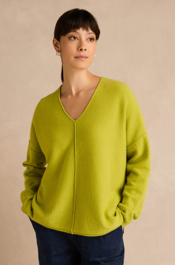 A woman wearing the Kiwi Merino Wool V Neck Jumper in lime green and dark blue pants stands against a beige background with her hands in her pockets.