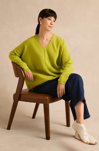 A person sits on a wooden chair, wearing the Kiwi Merino Wool V Neck Jumper in lime green, paired with dark blue jeans, white sneakers, and pearl earrings. Their hair is tied back as they pose against a neutral background.