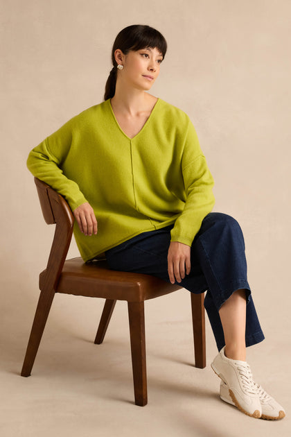 A person sits on a wooden chair, wearing the Kiwi Merino Wool V Neck Jumper in lime green, paired with dark blue jeans, white sneakers, and pearl earrings. Their hair is tied back as they pose against a neutral background.