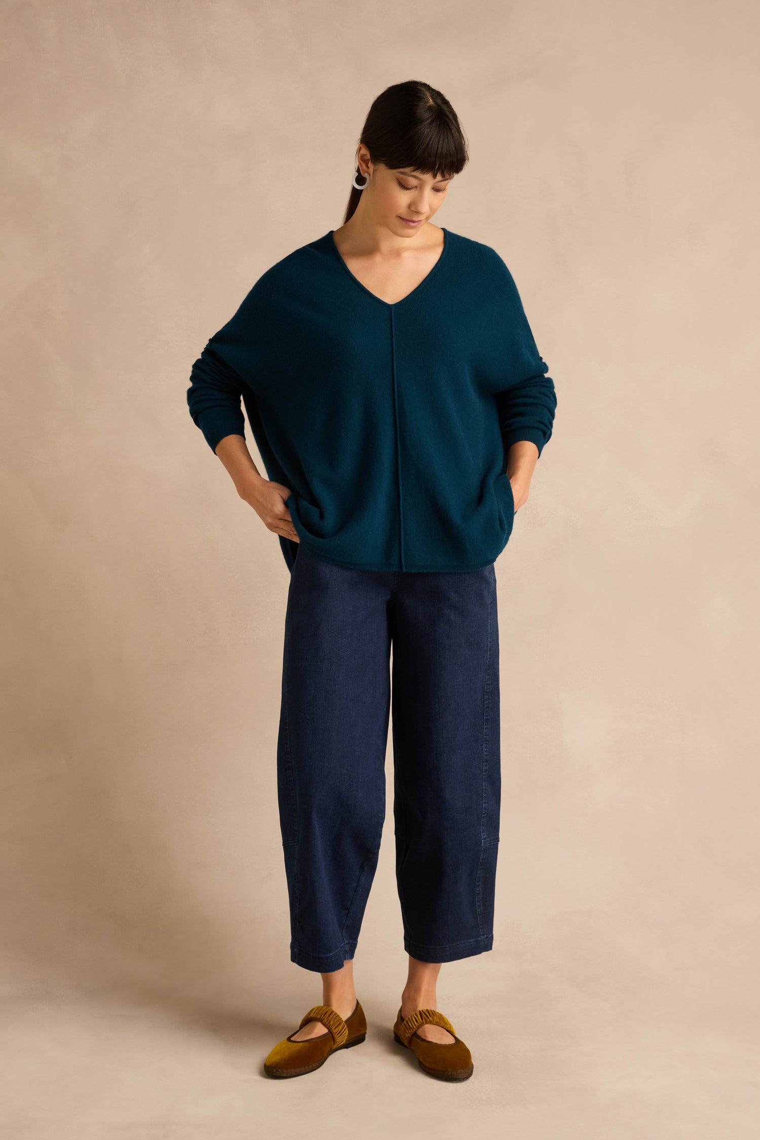 A woman stands against a beige background, wearing the Marine Merino Wool V Neck Jumper in teal, paired with dark blue wide-leg pants and brown loafers, her hands in her pockets as she looks down.