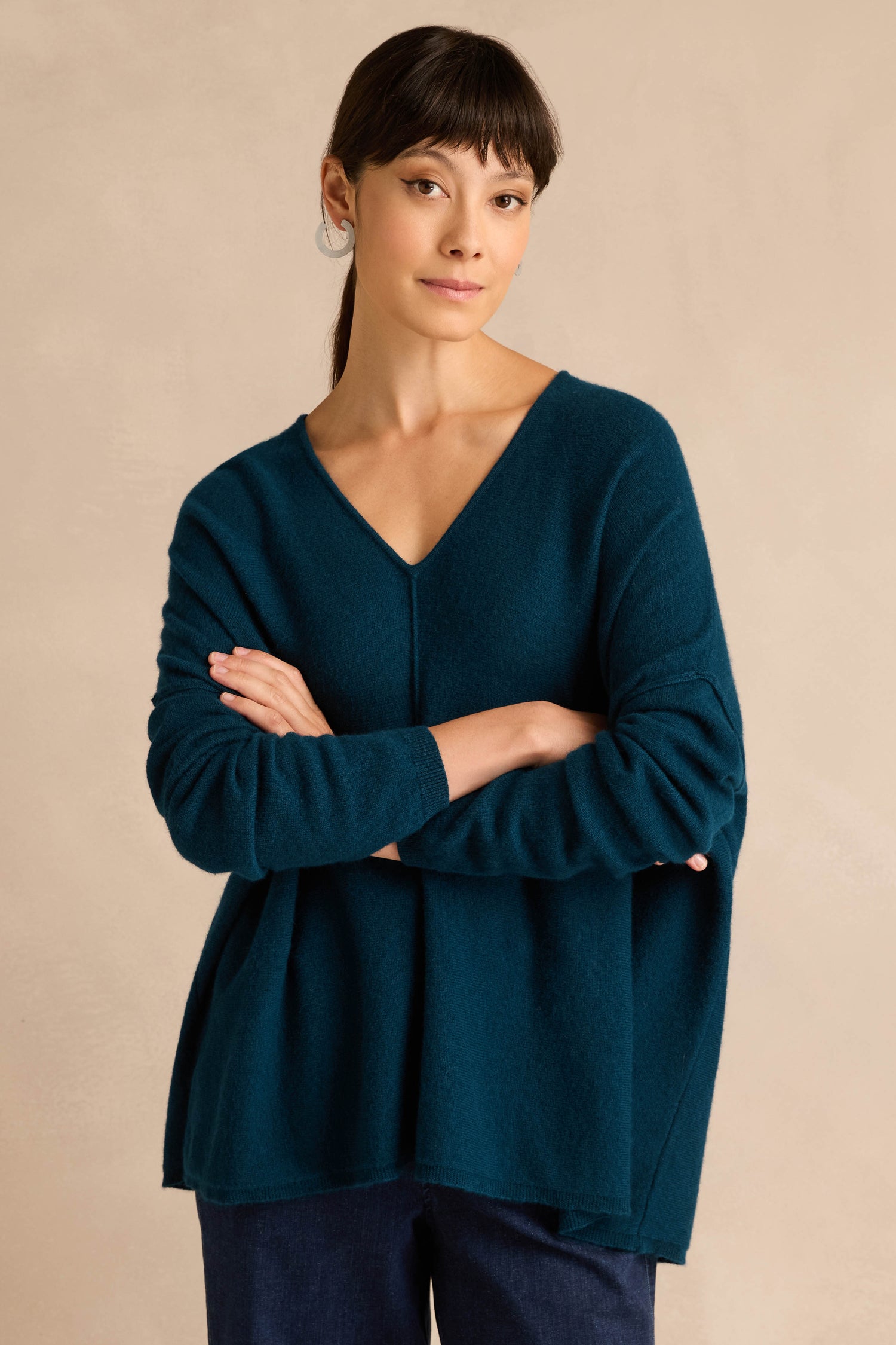 A woman with straight dark hair and bangs wears the Marine Merino Wool V Neck Jumper in a loose fit, paired with dark pants, standing with her arms crossed against a plain beige background.
