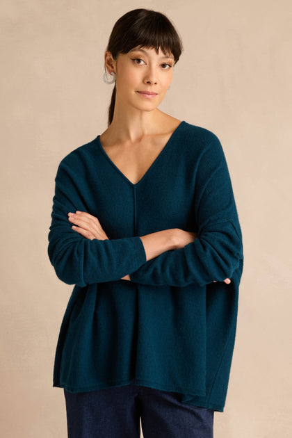 A woman with straight dark hair and bangs wears the Marine Merino Wool V Neck Jumper in a loose fit, paired with dark pants, standing with her arms crossed against a plain beige background.