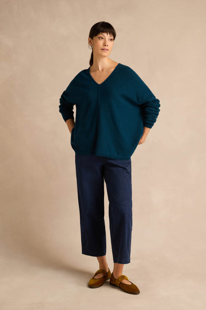 A woman stands against a beige background, wearing the Marine Merino Wool V Neck Jumper in teal, paired with dark blue cropped pants and brown shoes, with her hands in her pockets.