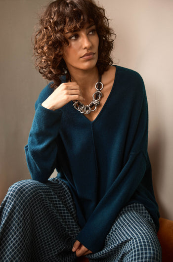 A woman with curly brown hair sits and looks to the side, wearing the Marine Merino Wool V Neck Jumper in dark teal, paired with patterned pants and a chunky silver necklace.